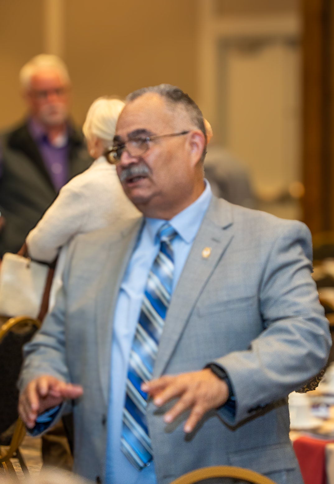 Community Builders Breakfast honored guest speaker and one of Habitat's latest new home owners, Luis Saldivar. Latin American male, greying hair, grey mustache, glasses, light blue suit jacket, shirt and tie.